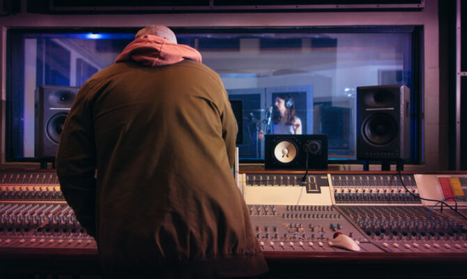 Sound engineer working at audio control panel with singer singing in recording room in background. Musicians producing music in professional recording studio.